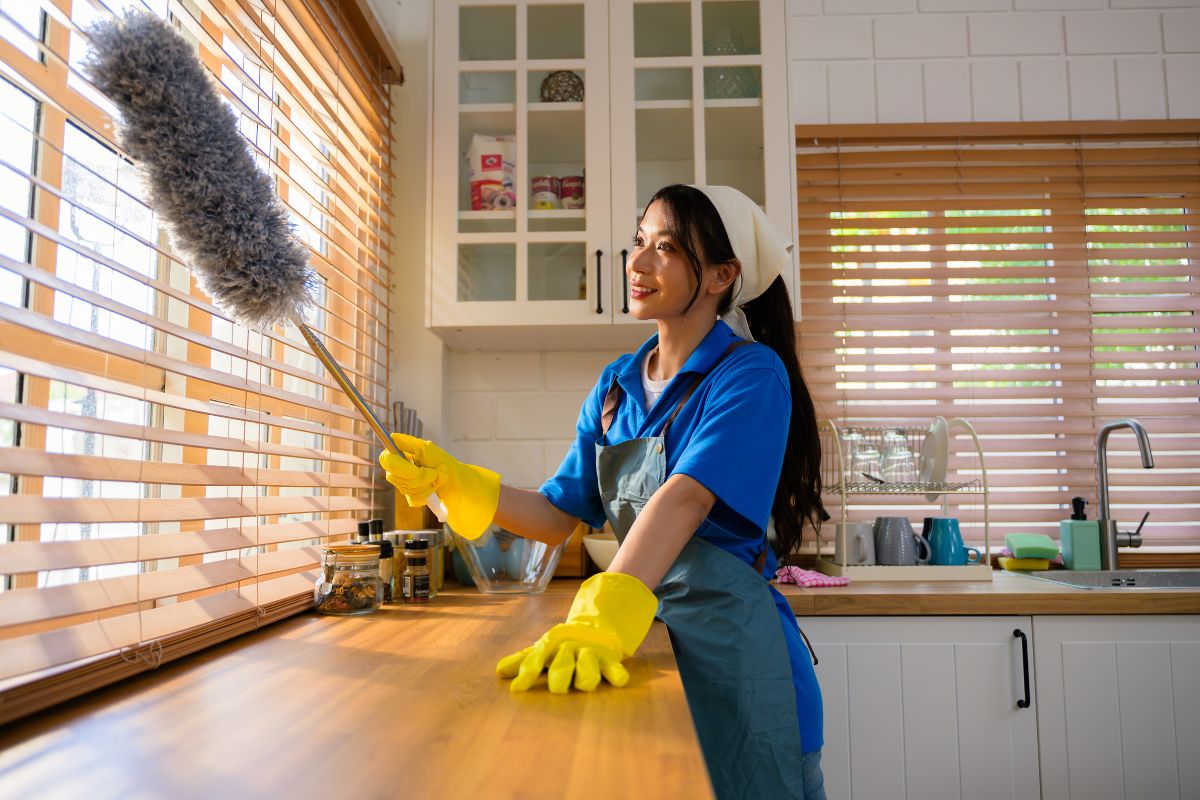 A house cleaner leaning against a wood counter and dusting window shades